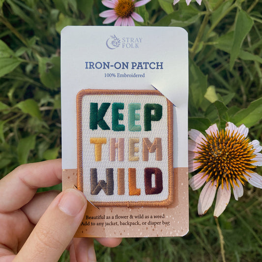 Colorful Keep Them Wild Iron-on patch, 100% embroidered, held over coneflowers. Packaging reads "Add to any jacket, backpack, or diaper bag."