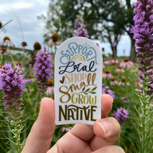 Sticker by Stray Folk being held in front of colorful native flowers. Sticker reads "Support Local, Shop Small, Grow Native" in various hand-drawn lettering styles with small illustration embellishments.