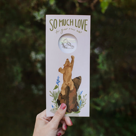 Hand holding a #10 baby card from the Stray Folk SunSpeak Series. In warm tones of pink, green, and browns, the greeting card depicts a painting of small cub standing on mother bear’s back, reaching upwards towards an iridescent suncatcher sticker that shows through a hole cut in the front. The suncatcher sticker has a line drawing of a mom bear with a cub on her belly. Around the bears are illustrated flowers and leaves. At the top of the card is the phrase "So much love for your new cub."