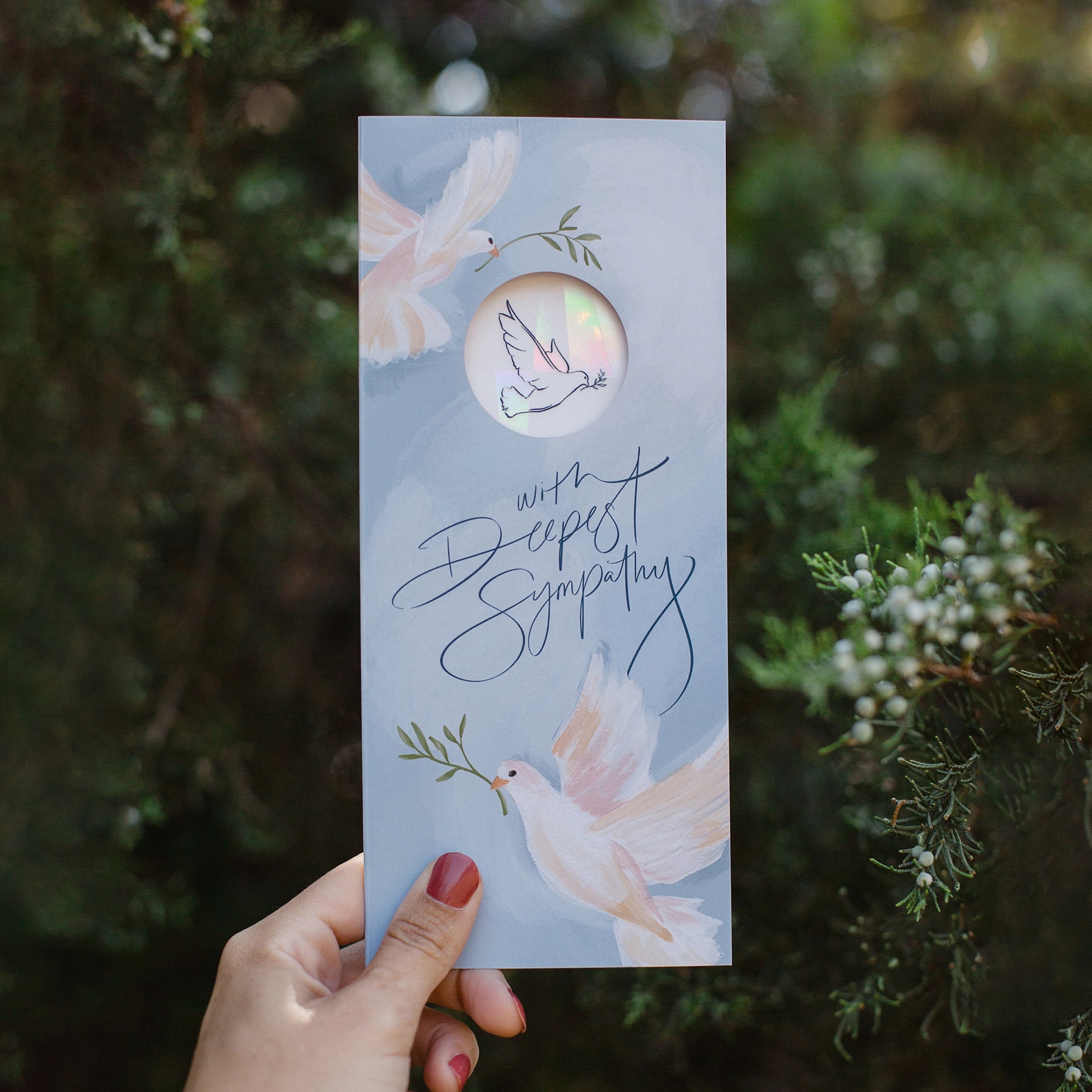 Hand holding a #10 sympathy card from the Stray Folk SunSpeak Series. In soft blues, creams, and pinks, the greeting card depicts two illustrated doves and the phrase “With Deepest Sympathy.” There is a hole cut out of the front where you can see the suncatcher sticker that's included inside. The suncatcher sticker is iridescent in the light and has small line drawing of a dove in the center.