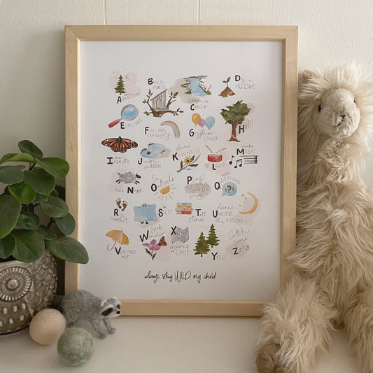 Framed ABCs of Childhood Alphabet Art Print by Stray Folk sitting on nursery table with stuffed animal and other whimsical decor