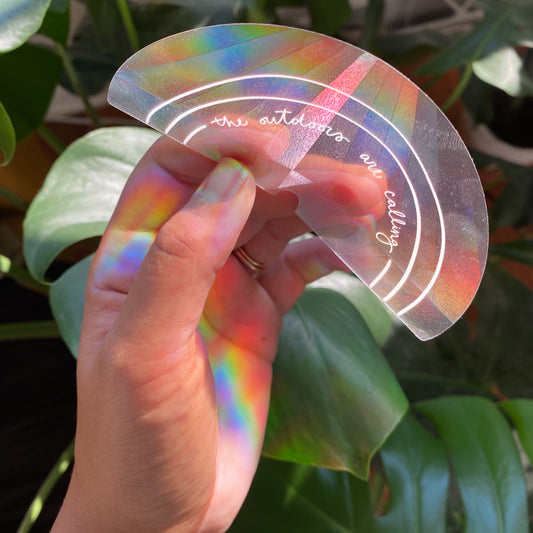 Hand holding transparent suncatcher sticker that says "the outdoors are calling" in front of plants to show rainbow effect from sunlight