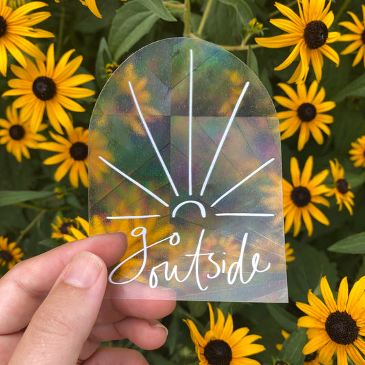 Transparent suncatcher sticker with words "go outside" held in front of wildflowers to show rainbow effect